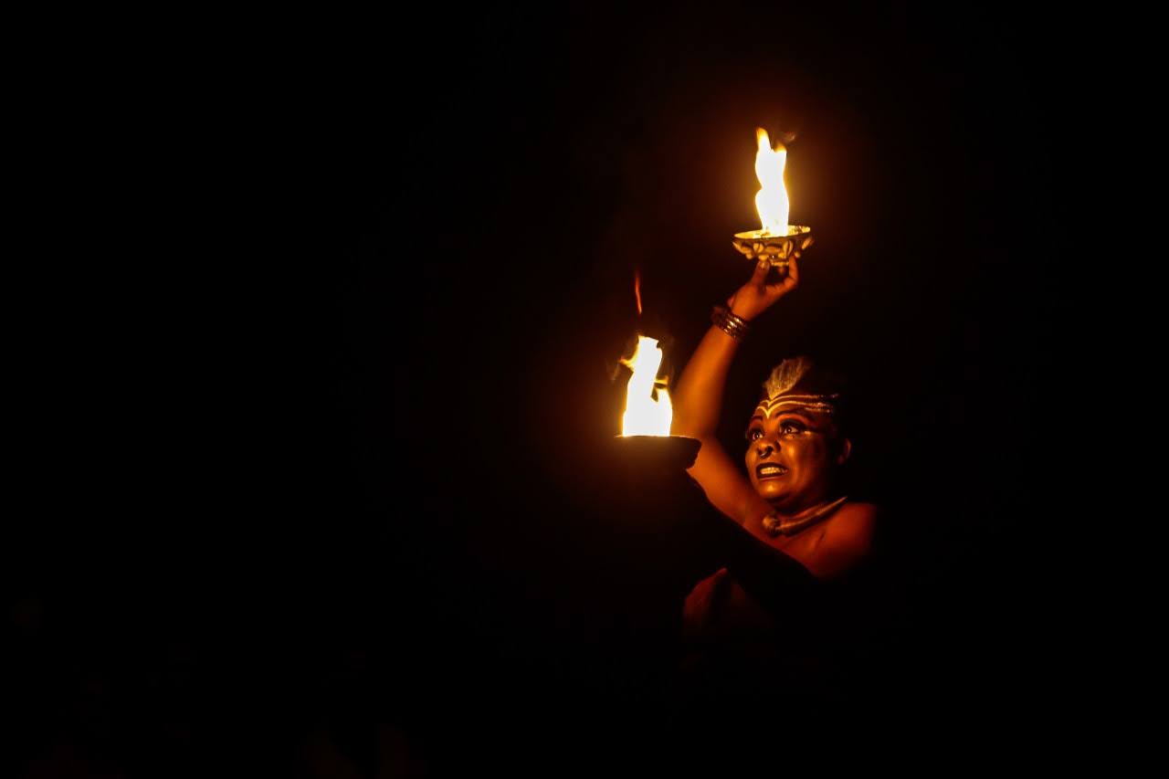 Márcia Limma looking at a flame, with her right arm raised, her face illuminated by the flame.