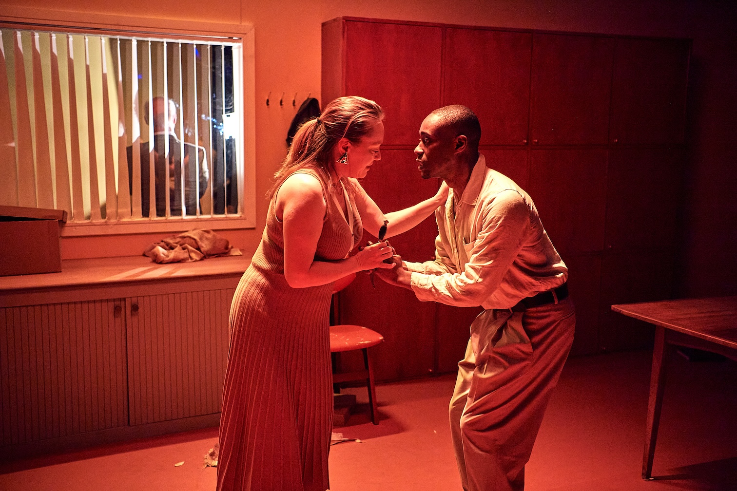 Actors playing Queen Merope [Rosie Thomson] and Aeyptus [Akiel Dowe] face each other, engaged in close conversation. Red light suffuses the space, which looks like an office interior.