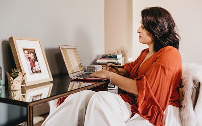 Female with dark hair and an orange top working at her desk using her laptop. She is surrounded by photos of her family, ornaments and books