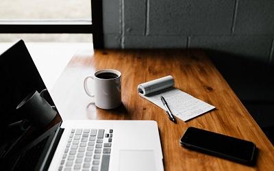 Image shows a laptop on a wooden desk next to a notepad and a cup of coffee. Photo credit Andrew Neel on Unsplash