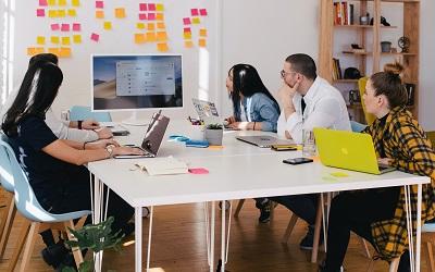 A team sit around a desk looking at a screen. They are in a meeting 