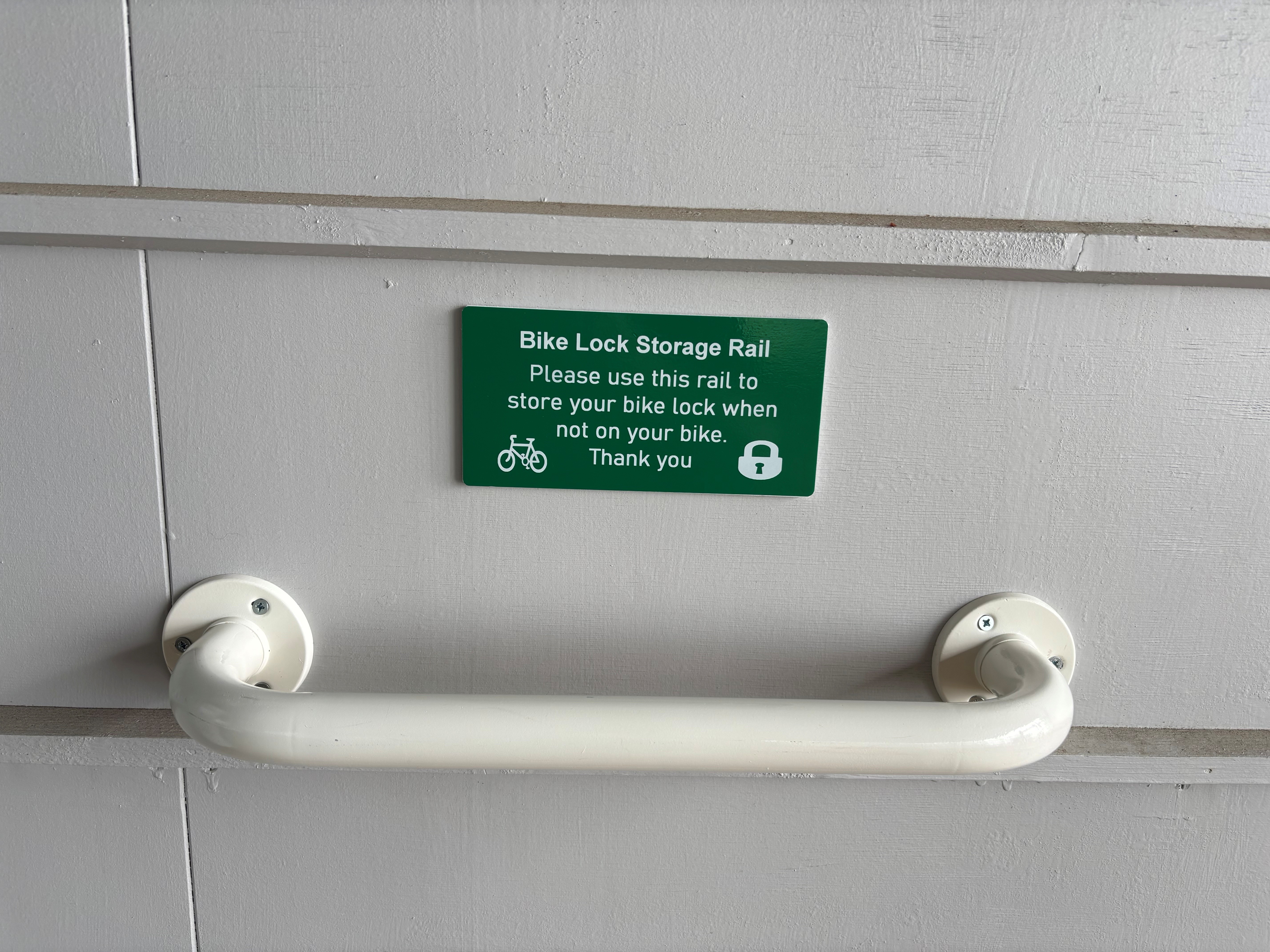 White storage rail mounted on a light grey wall in a bike shed, with a green sign above reading: “Bike Lock Storage Rail – Please use this rail to store your bike lock when not on your bike. Thank you.” The sign includes bicycle and padlock icons.