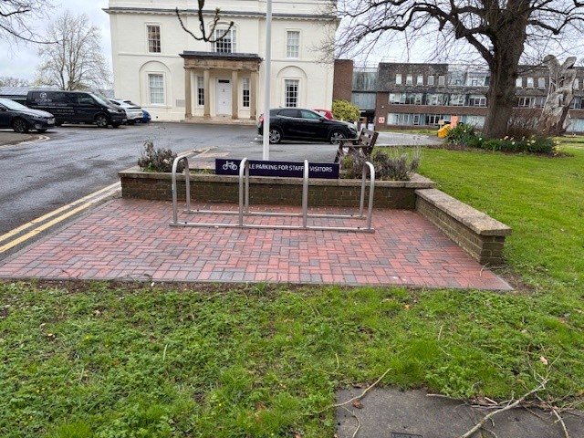 Outdoor cycle parking area with metal bike racks on a red-brick paved section, bordered by low brick walls, with a dark blue sign reading ‘Cycle parking for staff & visitors’ in front of a car park, with a white building and surrounding grass.