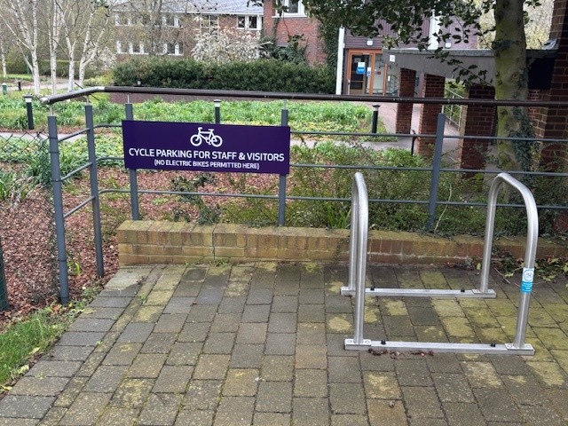 Outdoor cycle parking area with a metal bike rack and a dark blue sign reading ‘Cycle parking for staff & visitors (no electric bikes permitted here)’, located on a paved path.