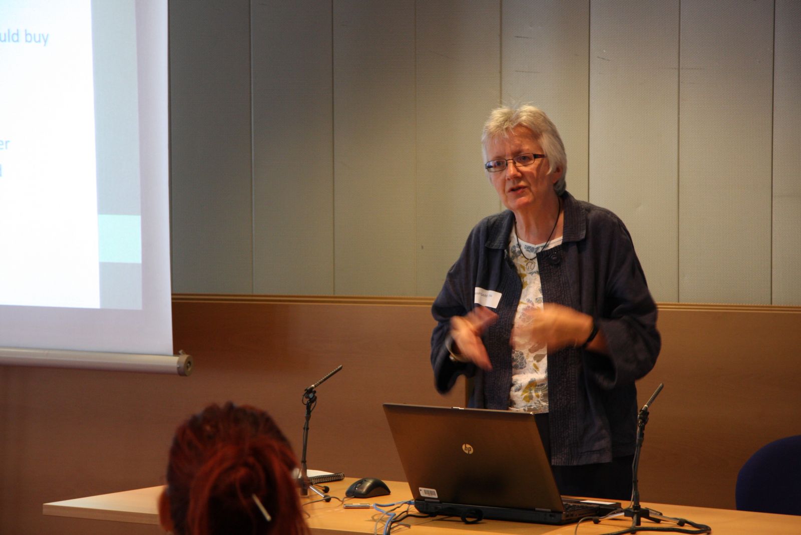 An image of a woman, standing behind a presentation table, giving a speech