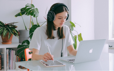 A woman with brown hair and headphones on writing on an ipad to her right while working on a laptop.