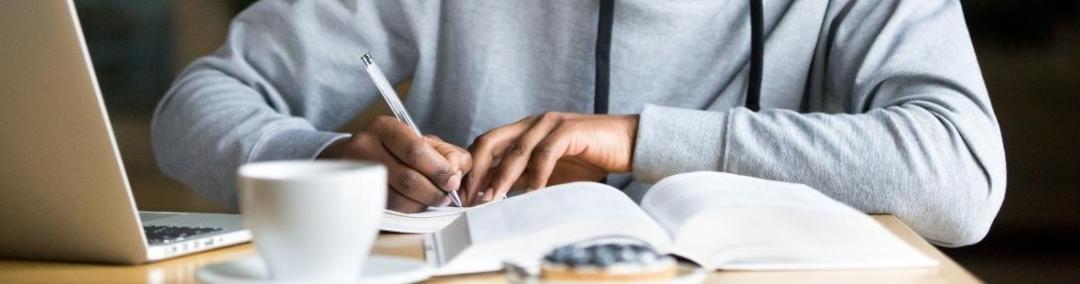 a young man writes in a notebook at a desk with a text book in front of him