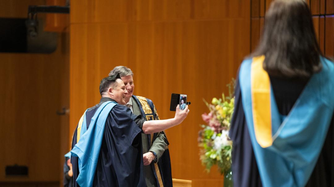 A student during the OU Graduation ceremony. He is on stage in his OU robe, taking a selfie with the Presiding Officer. They are both smiling.