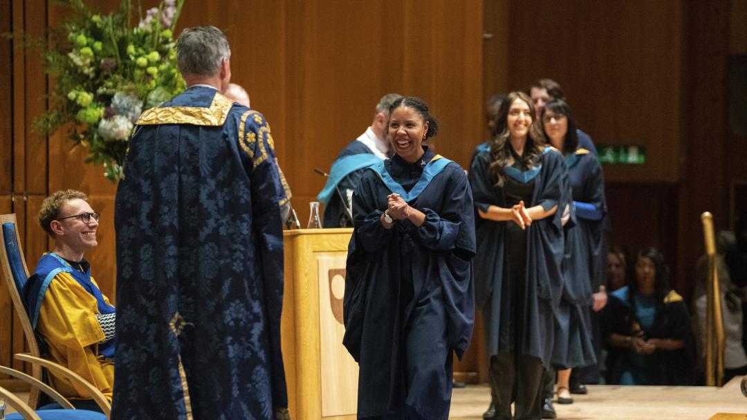 A student during the OU graduation day. She is wearing the OU robe and walking across the stage. She is smiling joyfully.