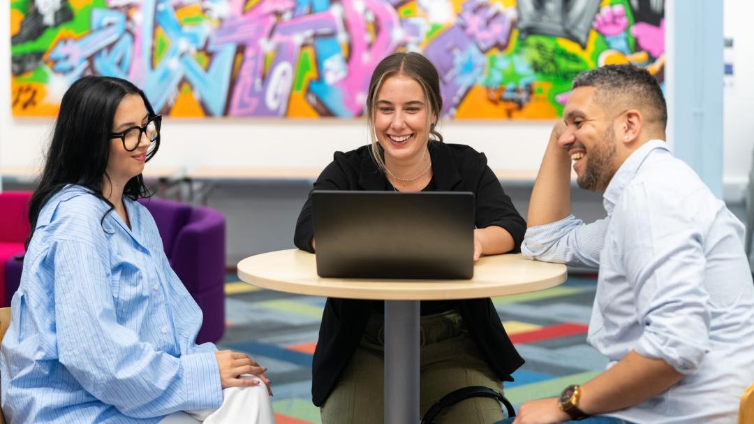 Three students sitting around a cafe table. They are smiling and checking something on a laptop.