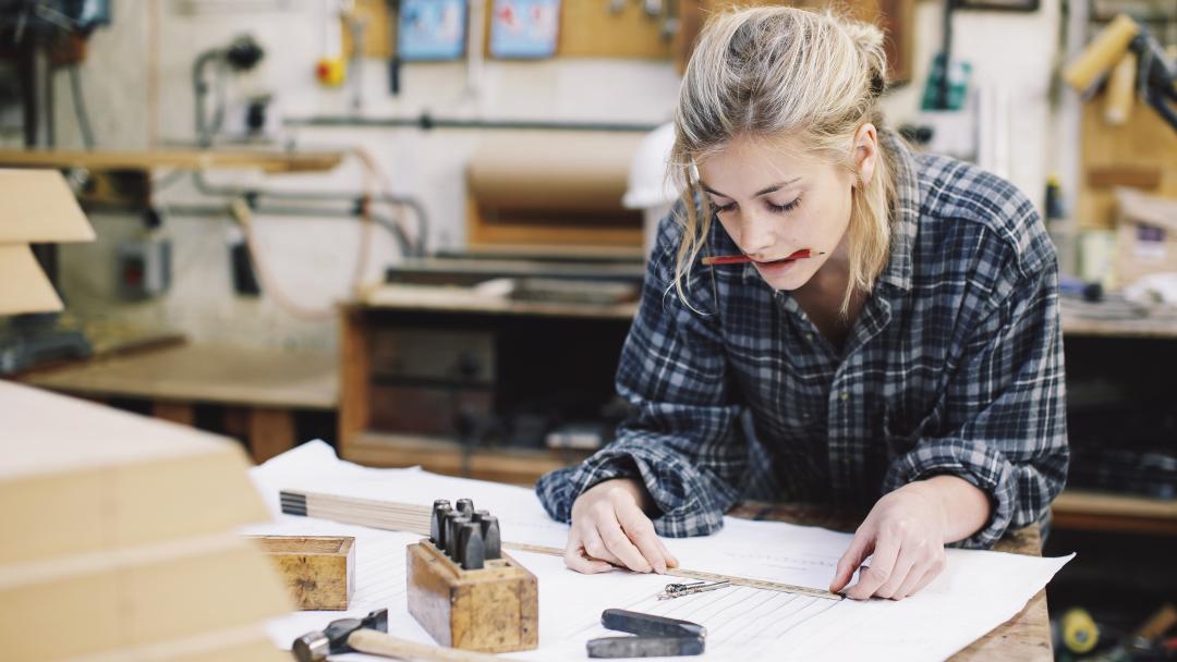 An engineering student. She is leaning over a workbench, making measurements on a sheet of paper. She is focused.