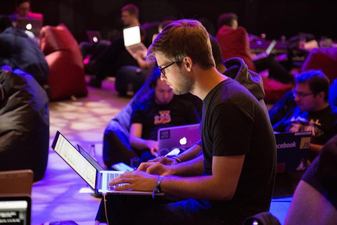 A student sitting on the floor of a conference room. He is working on his laptop. There are other people sitting around him, also on their laptops.