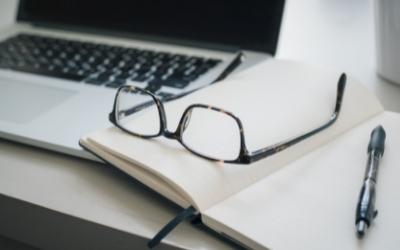 A laptop on a desk with a notebook beside it and a pair of glasses and a pen on top of it