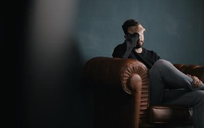 A man looking sad on a leather brown sofa in a dark room with his hand over his face