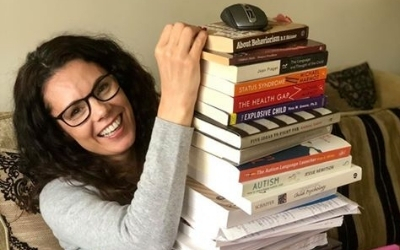 A female student smiling while sitting on the sofa with a large pile of text books on her lap