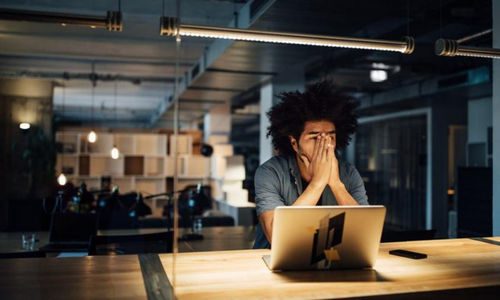 A man sitting at a desk with hands over his face looking worried in front of a laptop