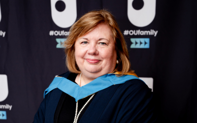 Anna smiling, in academic robes, standing in front of a screen printed with OU logos and #OUFamily. 