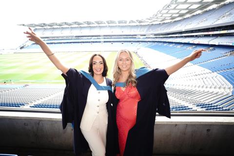 Two women wearing graduation gowns and smiling joyfully with their arms raised, standing in front of empty stands at a large sports stadium, likely celebrating their graduation. The background shows a green pitch and rows of blue and white seats.