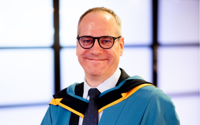 Paul smiling, in blue academic robes, standing in front of a screen.