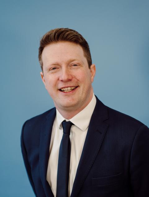 “Professional head-and-shoulders portrait of a man wearing a navy suit, white shirt and dark tie, smiling slightly against a plain light blue background.”