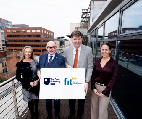 Four professionally dressed individuals stand on a modern outdoor balcony in an urban setting, holding a sign featuring the logos of The Open University in Ireland and FIT (Fast Track to IT), an organization focused on tech talent development. The group consists of two men and two women, all smiling at the camera. The man in the center, wearing a light grey suit and orange tie, and the bald man to his left in a navy plaid suit, hold the sign together. The background features modern office buildings with lar