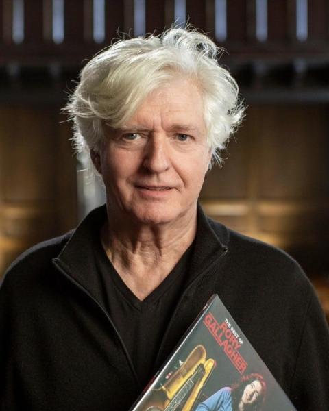 “Older man with white hair wearing a black top, holding a book titled The Best of Rory Gallagher with a guitar image on the cover, standing in a softly lit indoor setting.”