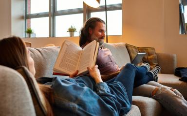 Couple relaxing on a couch in a sunlit room, one reading a book while the other uses a tablet.