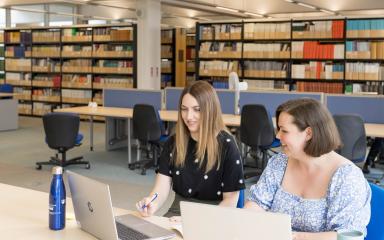 Two people are sitting next to each other at a desk in the Library building, looking at open laptops. Behind them are banks of desks and rows of bookshelves.
