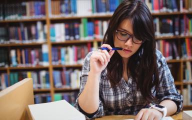 A student is sitting at a desk in a Library with a pen to her mouth, thinking about what to write. Behind her is a large bookshelf.