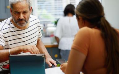 A mature student is looking at the screen of a tablet computer which is sitting on a kitchen counter. He is surrounded by family members.