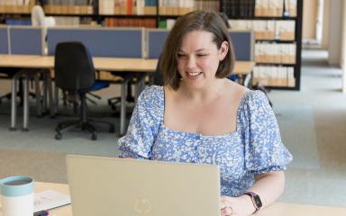 A woman in a blue floral dress is working on a laptop in a quiet library with shelves of books behind her.