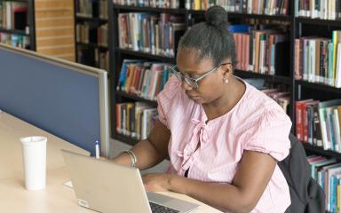 A student sitting at a desk writing in a notepad next to an open laptop. There is a row of bookshelves behind them.