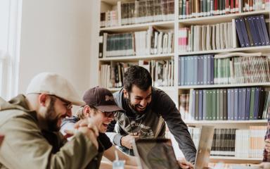 A group of students are sitting round a table looking at an open laptop and laughing. There is a shelf of books behind them