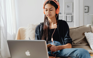 A student wearing headphones is sitting on a sofa writing in a notepad, with an open laptop in front of them.