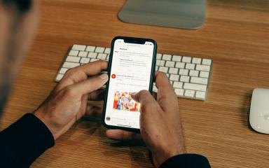 Person scrolling through social media feed on smartphone at wooden desk with keyboard and mouse nearby.