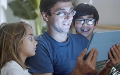 A family are sitting on a sofa together looking at a tablet computer whilst smiling