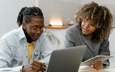 Two people are sitting next to each other at a desk. One is holding a tablet computer and resting their hand on a book. The other is typing on an open laptop.