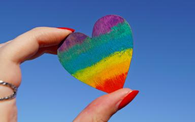 Close-up of a hand holding a multicoloured heart ornament, representing LGBT+ pride colours.