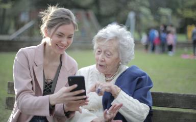 Young woman showing something on her smartphone to an elderly woman sitting together on a park bench during daytime.