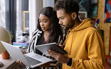 Two students in a home environment are looking at an open laptop, with one student holding a tablet computer