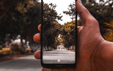 Close-up of a person's hand, holding a phone whilst taking a photo of a long road surrounded by trees