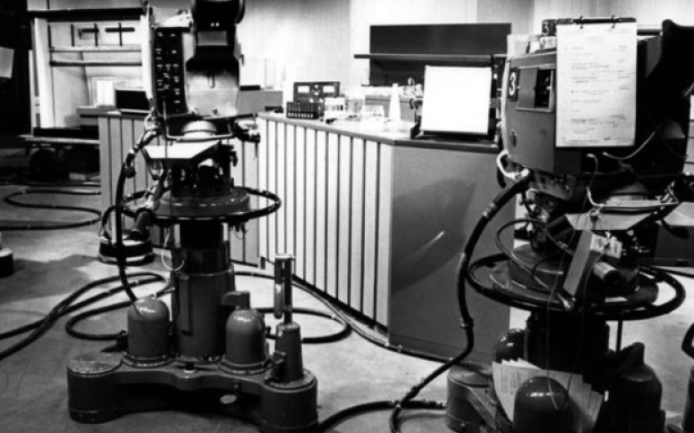 A black and white photo of a television recording studio, featuring large broadcast camera equipment.