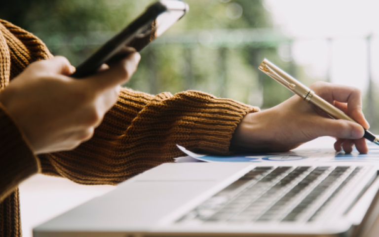 Close-up of a person's hands, holding a phone in one hand and a pen in the other. An open laptop is also present.