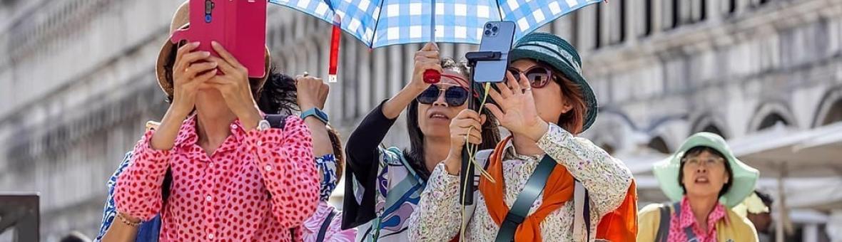Asian tourists taking photos with their mobiles. One woman is holding an umbrella over the two women who take the photos. 