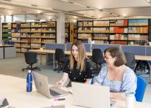 Two people are sitting next to each other at a desk in the Library building, looking at open laptops. Behind them are banks of desks and rows of bookshelves.