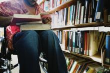 A student in a wheelchair has a pile of books resting in their lap. A stack of books can be seen behind them.