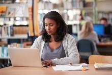 A student is sitting at a desk in a Library, looking at an open laptop