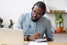 A student is sitting at a desk, studying at home on his laptop. He is wearing headphones and writing notes.