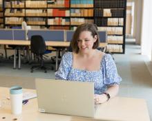 A woman in a blue floral dress is working on a laptop in a quiet library with shelves of books behind her.