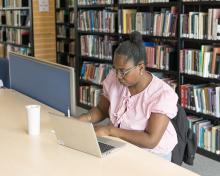 A student sitting at a desk writing in a notepad next to an open laptop. There is a row of bookshelves behind them.